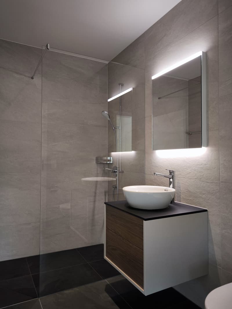 Modern bathroom with a glass shower enclosure, a white sink, a mirror with LED lighting, and a wooden cabinet.