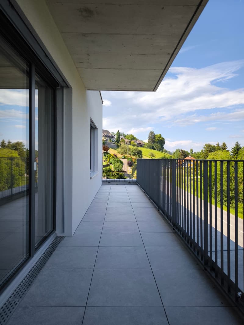 A balcony with a tiled floor, glass railings, and a view of a hillside with houses and trees.