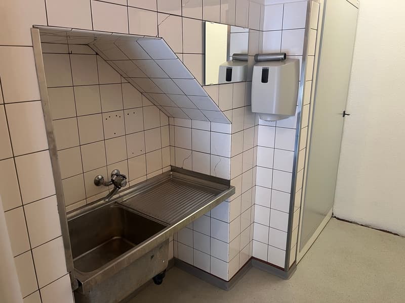 Room with white tiled walls, metal sink with metal tray, a mirror, and a soap dispenser
