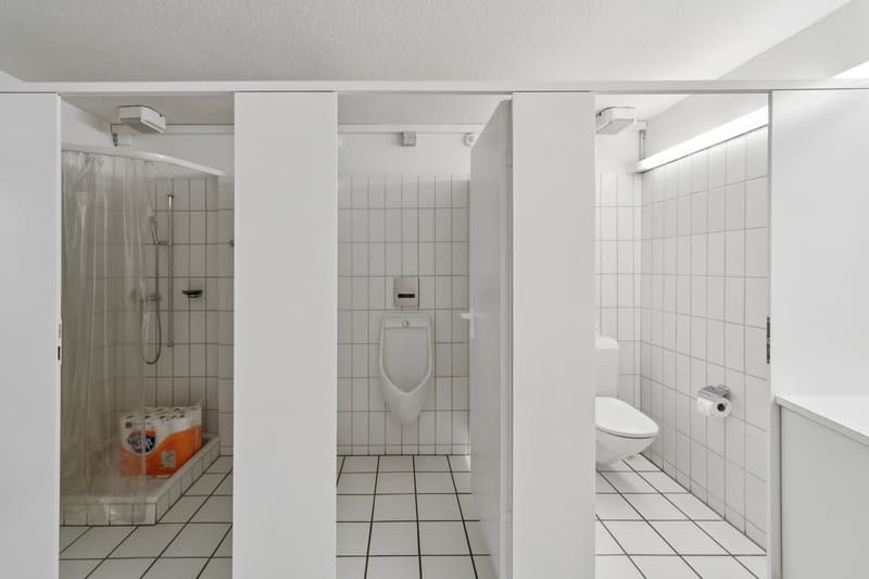 A bathroom with white tiles, a shower, a urinal, a toilet, and a toilet paper holder.