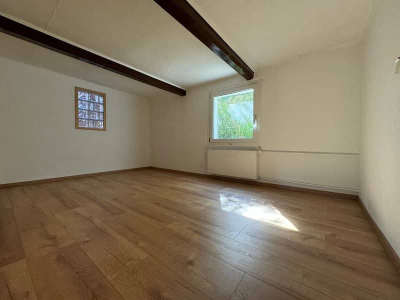 This image shows an empty room with hardwood floors, white walls, and exposed wooden beams on the ceiling. There is a small window that provides natural light to the space.