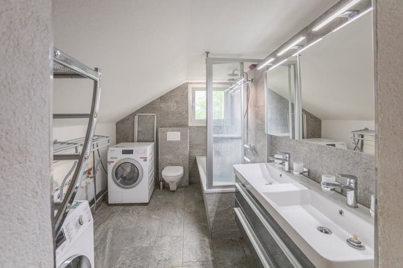 Spacious bathroom with gray tile flooring, a washing machine, a dishwasher, a vanity with a sink, and a glass-enclosed shower area