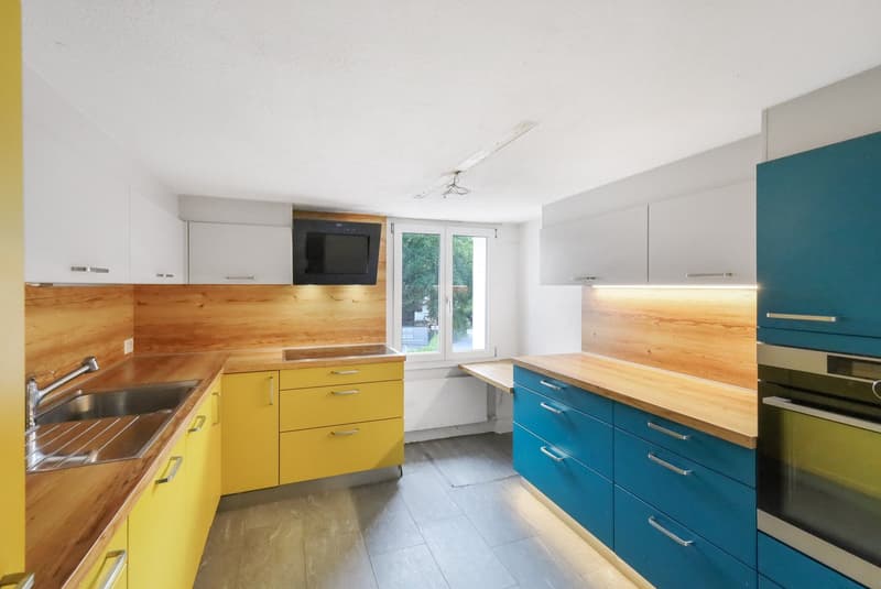 Modern kitchen with wooden countertops, yellow and blue cabinets, stainless steel sink, and a built-in dishwasher. The kitchen has large windows providing natural light.