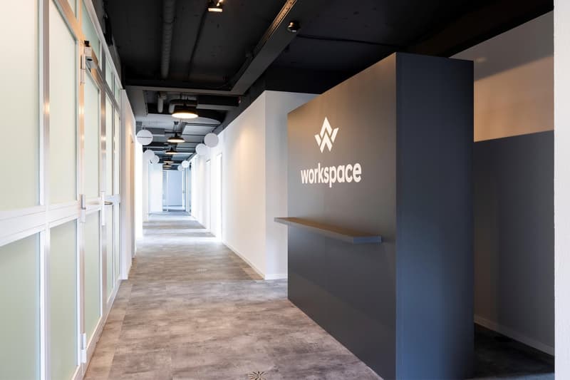 The image shows a modern and minimalist office space. The hallway has a concrete floor, white walls, and a black ceiling with exposed pipes and lighting fixtures. The Workspace logo is prominently displayed on the wall, indicating this is a shared workspa