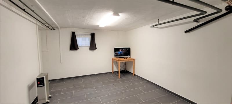 small room with white walls, gray tiles, a table with a monitor, a heater, and a window with curtains