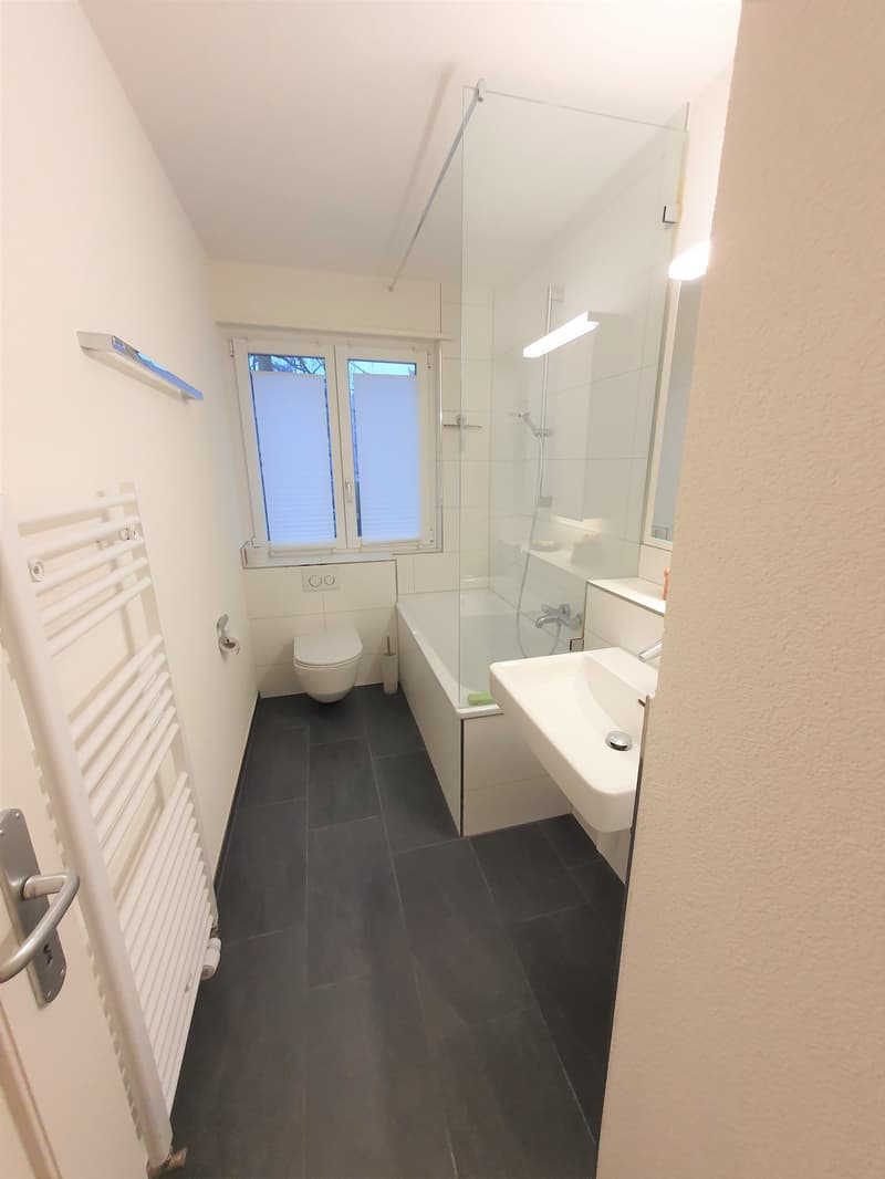 Bathroom with white tiles, glass shower enclosure, white sink, white toilet, towel rack, and a window.