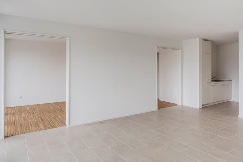 Empty room with white walls, wooden floor, and tiled floor, kitchen area with sink and cabinets