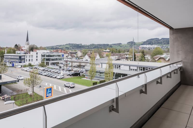 Balcony with a view of the city, metal railing, tiled floor, cars parked in the parking lot, trees, and buildings in the distance