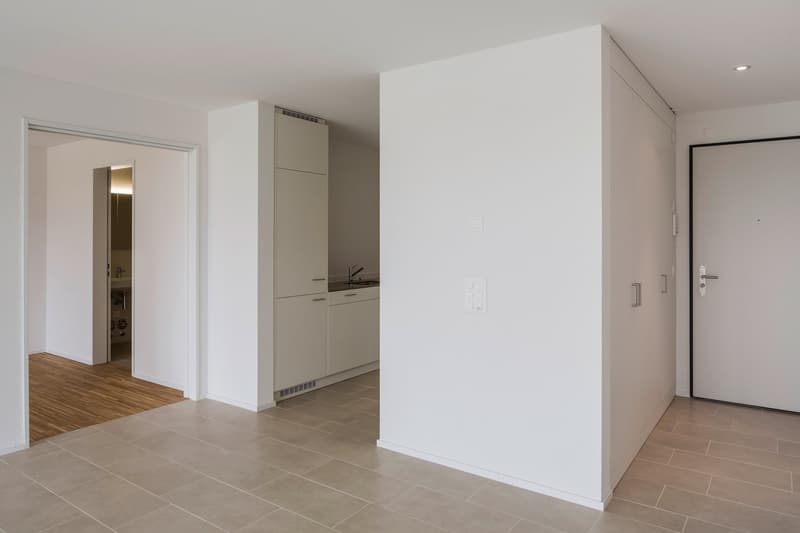 Empty room with white walls, tiled floor, kitchen area with sink and cabinets, entrance door, and a door leading to another room