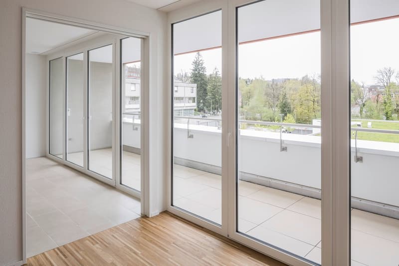large sliding glass doors, wooden floor, white tiles, balcony with railing, view of trees and buildings