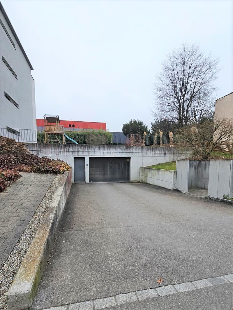 An empty parking lot with concrete walls and a garage entrance. The area is surrounded by buildings and has a small tree and a slide in the background.