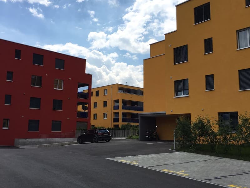 Two multi-story apartment buildings, one red and one yellow, concrete parking area, metal railings, green vegetation, clear blue sky with clouds