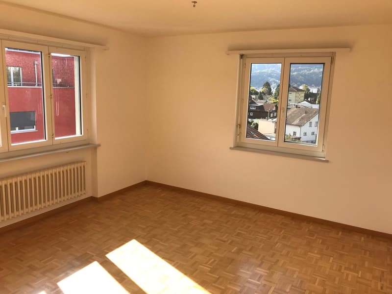 Spacious room with hardwood flooring, large windows providing natural light, and a view of the surrounding buildings and landscape.
