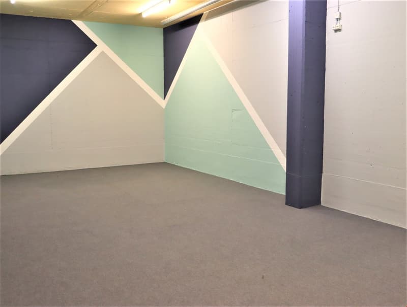 This image shows a large, open room with a concrete floor and painted walls. The walls are painted in a combination of dark blue and light mint green, creating a geometric pattern. There is a concrete column visible in the corner of the room. The room app