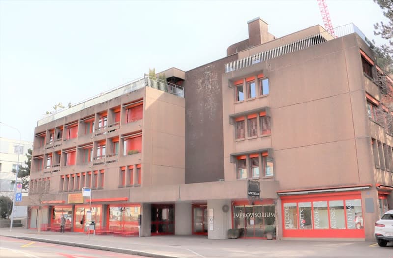 Multi-story commercial building with red accents, balconies, and retail spaces on the ground floor
