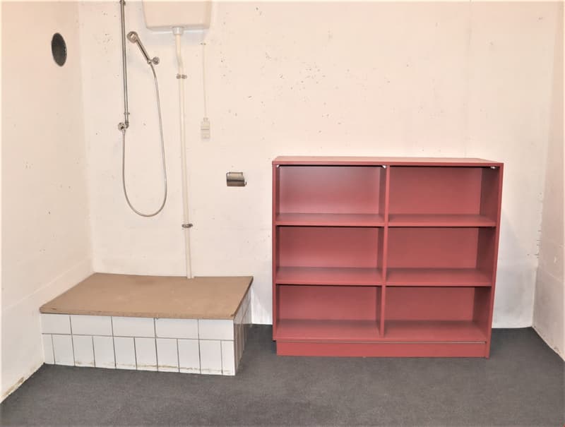 The image shows a bathroom with a shower stall and a red shelving unit. The floor is tiled and there is a raised platform, likely for a bathtub or other fixture.