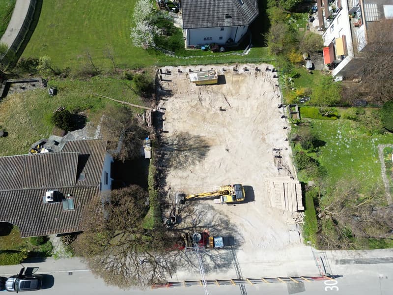 Aerial view of a construction site with a yellow excavator, a large open area, and several houses in the background.