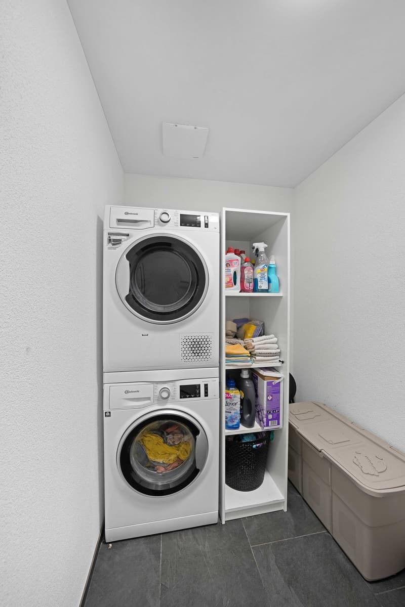 Laundry room with stacked washer and dryer, shelves with cleaning products, and a storage box.