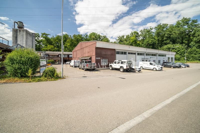 The image shows a commercial property with a parking lot in front. The building has a brown exterior and appears to be a warehouse or industrial facility. There are several vehicles parked in the lot, including work trucks and vans. The surrounding area i