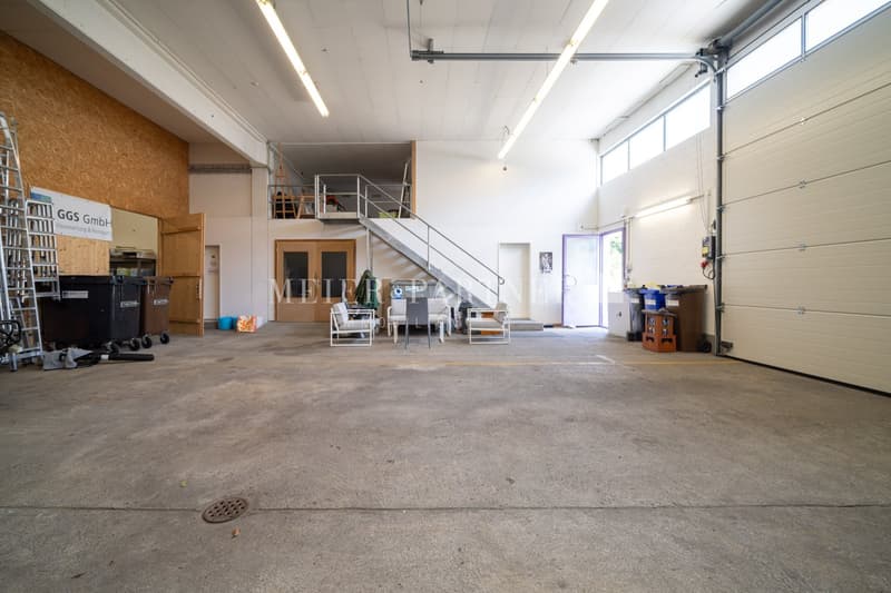 Large open space with concrete floor, metal shelving units, and a staircase leading to an upper level. The space appears to be used for storage or a warehouse, with the GGS GmbH company name visible on the wall.