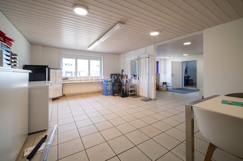 Large open space with tiled flooring, white walls, and a suspended ceiling with recessed lighting. The room contains various household appliances and storage items, including a washing machine and dishwasher.