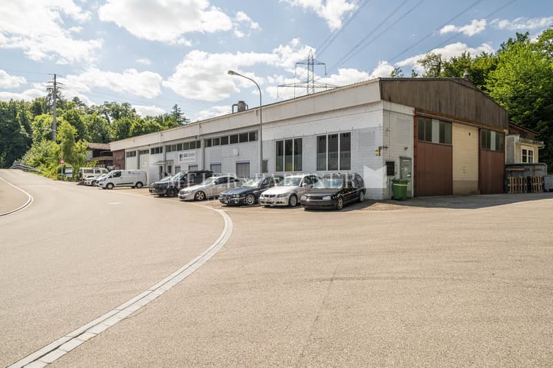 A commercial building with a parking lot in front. The building has a rectangular shape with large windows and a garage door. Several cars are parked in the lot. The surrounding area includes trees and power lines.