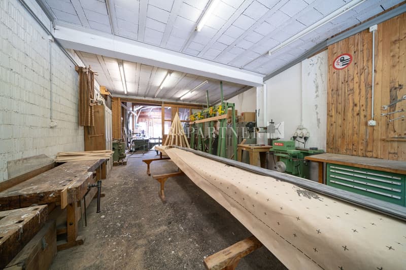 Large workshop space with wooden shelves, workbenches, and various tools and equipment. The ceiling is made of wooden beams and the walls are a mix of brick and wood paneling. There appears to be a garage door or entrance visible in the image.