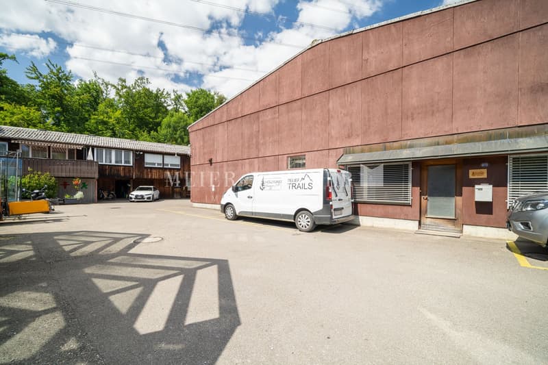 A commercial building with a large brown exterior wall, a delivery van parked in the front, and a paved parking area surrounded by trees and greenery.