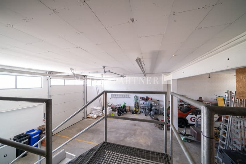 This image shows a spacious garage or workshop area. The space has a concrete floor, white walls, and a high ceiling with exposed beams. There are various tools, equipment, and storage shelves visible, indicating this is a functional workspace. A motorcyc