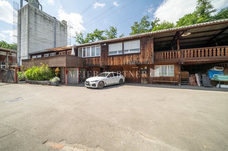 The image shows a commercial property with a wooden exterior and a balcony. There is a car parked in the front, indicating a parking space. The property appears to be a business or office space, with the 'Meyer & Partner' sign visible on the building.