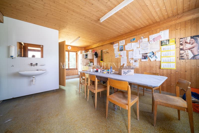 The image shows a rustic kitchen and dining area with a wooden ceiling and walls. There is a large table with chairs, a sink, and various items and calendars displayed on the walls.
