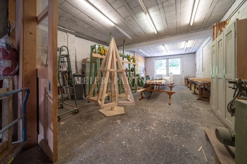 The image shows a workshop or studio space with a high ceiling, concrete floor, and various tools and equipment. There are wooden shelves, ladders, and a large wooden structure that appears to be a frame or sculpture. The space seems to be used for woodwo