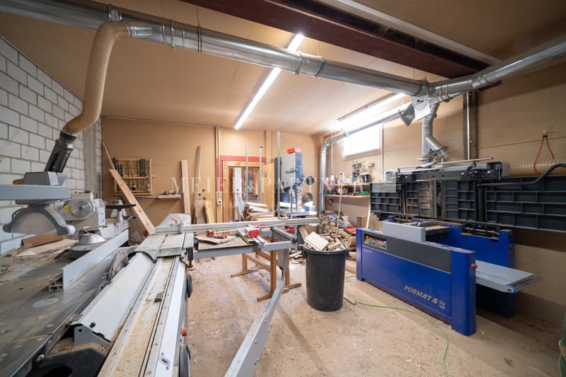 The image shows a workshop or garage-like space with various woodworking tools and equipment. There are power tools, workbenches, shelves, and storage containers visible. The space has exposed ductwork and a concrete floor, indicating an industrial or wor