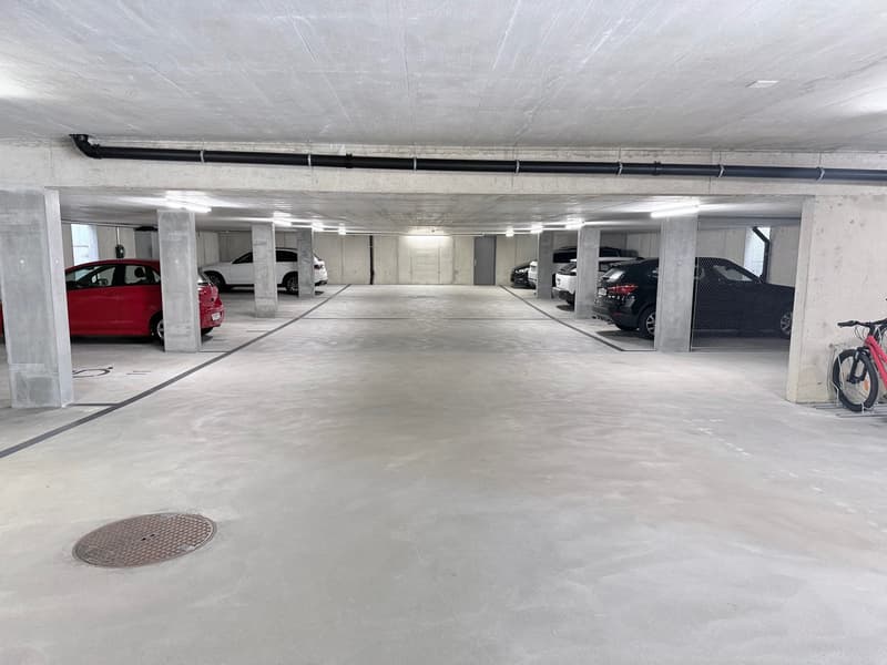Underground parking area with 5 cars parked, concrete floor, ceiling pipes, a manhole cover, and a bicycle rack.