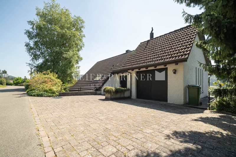 single story house, brown shingled roof, garage, brick driveway, green trash bin
