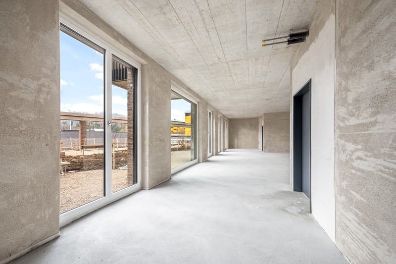 Long, wide hallway with concrete walls and ceiling, large windows, and a concrete floor. The hallway appears to be part of a commercial or industrial building under construction or renovation.