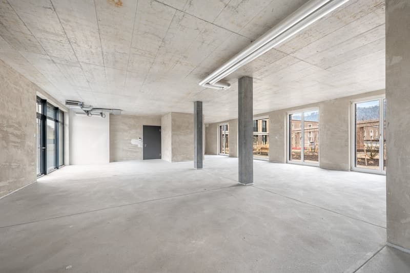 Large open-plan commercial space with concrete floors, exposed concrete ceiling, and large windows providing natural light. The space has a minimalist, industrial aesthetic with concrete columns and a door leading to a balcony or terrace area.
