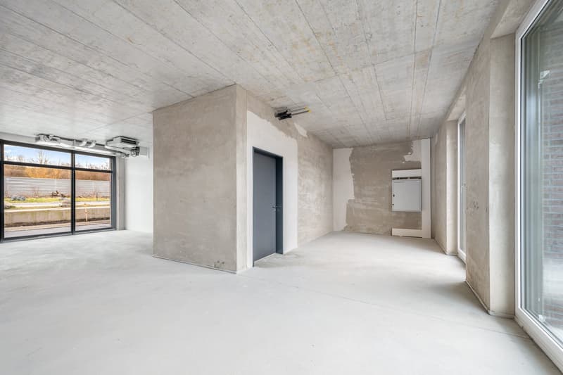 Large open space with concrete walls and ceiling, minimal furnishings, and a large window providing natural light.
