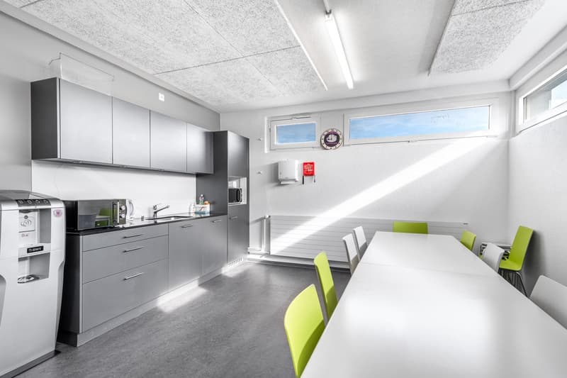 Gray cabinets, a sink with a faucet, a microwave on the counter, a coffee machine, a refrigerator, a desk with chairs, and a window.