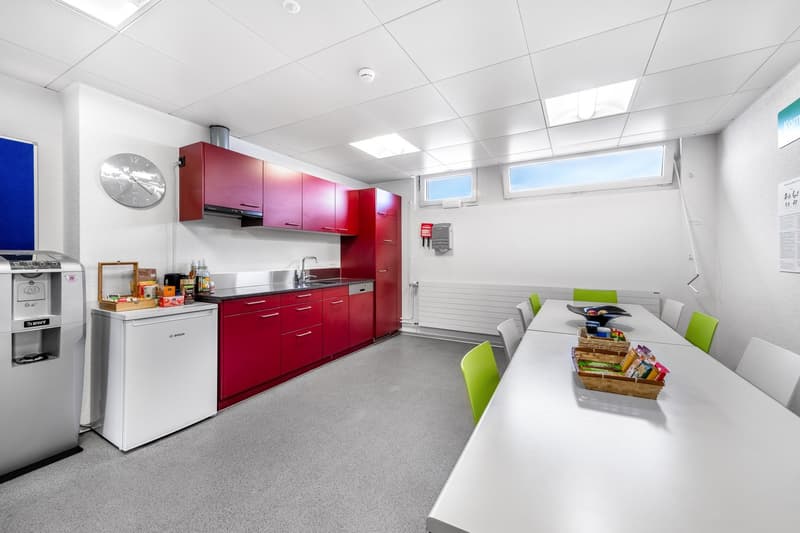 White walls, white ceiling, red cabinets, white sink, water dispenser, red wall clock, white dining table, green chairs, two windows on wall