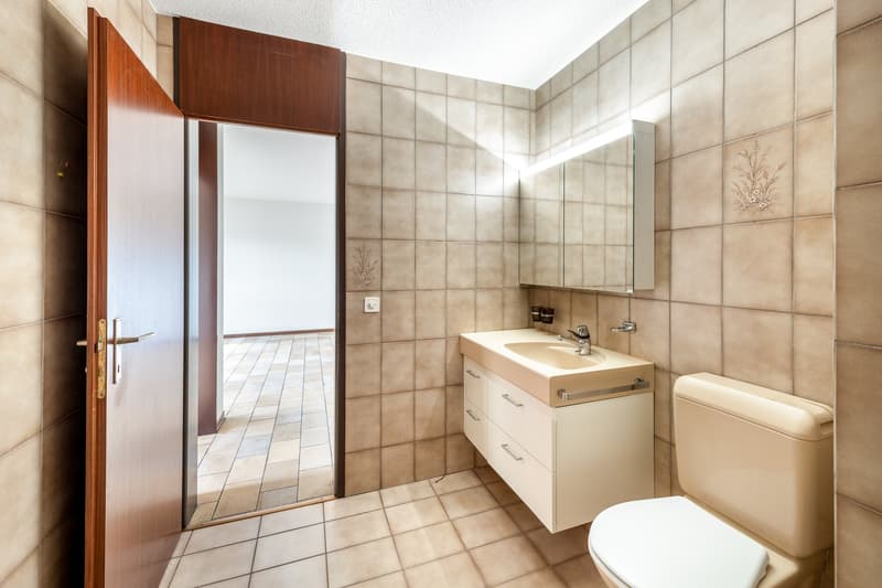 A small bathroom with beige tiles, a sink, a mirror, a toilet, and an open door leading to another room.