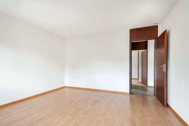 Empty room with white walls, wooden floors, and an open door leading to another room with a mirror.