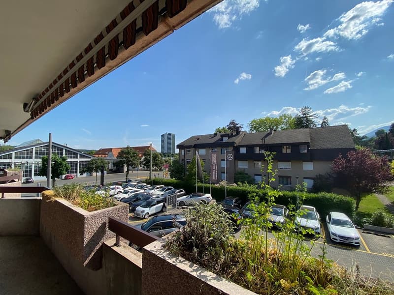 Parking lot with multiple cars, Mercedes-Benz dealership sign, greenery, trees, mountains in the distance, apartment building, cloudy sky