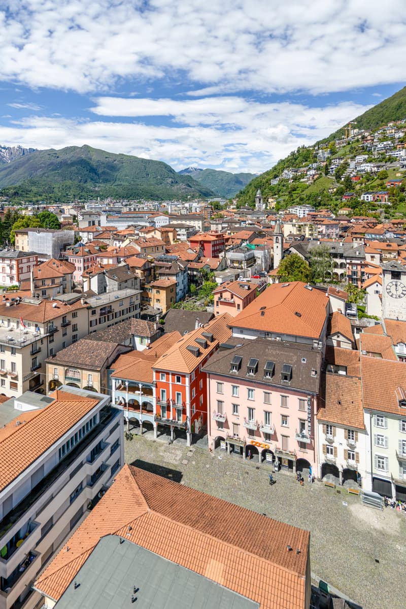 Aerial view of a picturesque city nestled in a mountainous landscape. The city features colorful buildings with red tile roofs, winding streets, and a church steeple. The surrounding mountains and blue sky with fluffy clouds create a scenic backdrop.