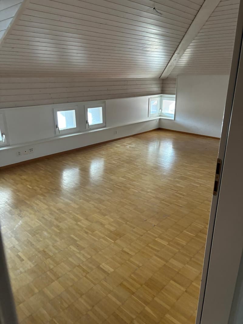 Spacious attic room with wooden paneled ceiling, hardwood flooring, and two small windows providing natural light.