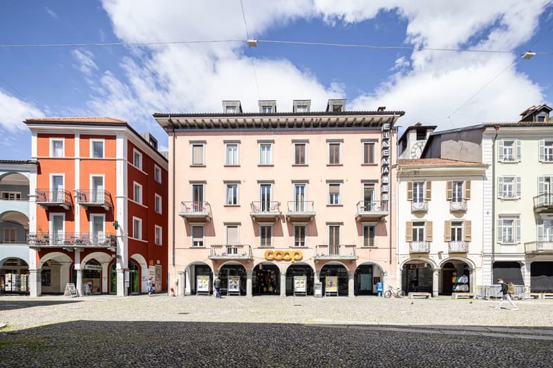 This image shows a row of multi-story buildings with arched entrances and balconies in a European-style town square. The buildings have a mix of orange, pink, and white facades, and there is a Coop supermarket visible on the ground floor of one of the bui