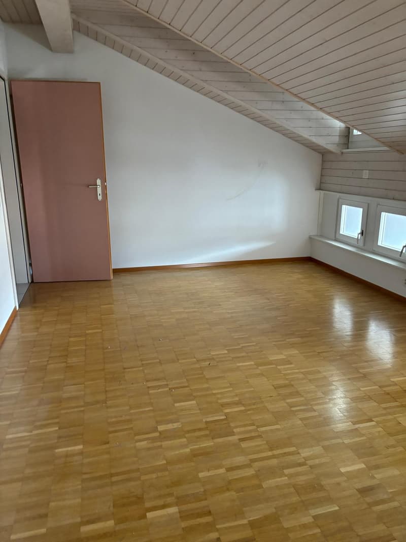 This image shows a spacious attic room with a slanted wooden ceiling and hardwood flooring. The room has a pink door and large windows, providing natural light. The walls are painted white, creating a bright and airy atmosphere.