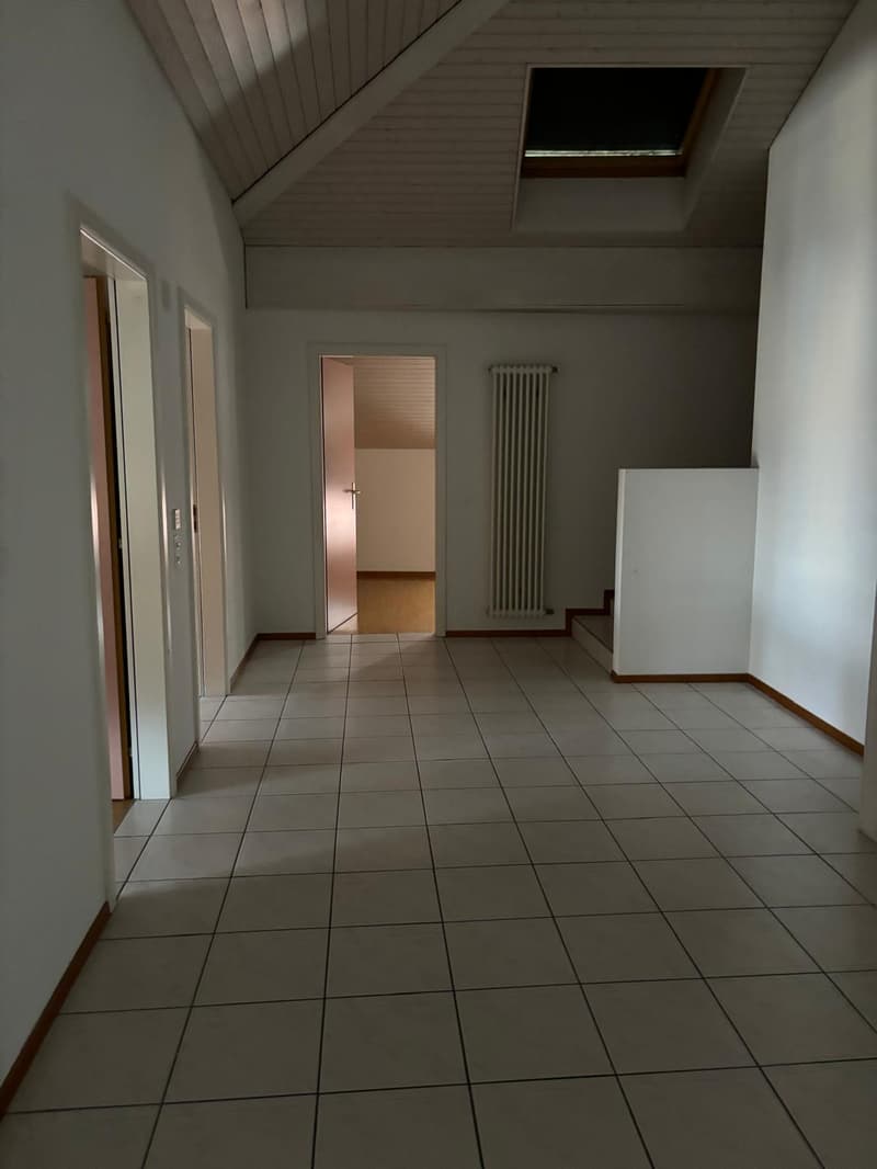 The image shows a room with a tiled floor, white walls, and a radiator. There are two doors visible, one leading to another room. The ceiling has a slanted design with a skylight window.