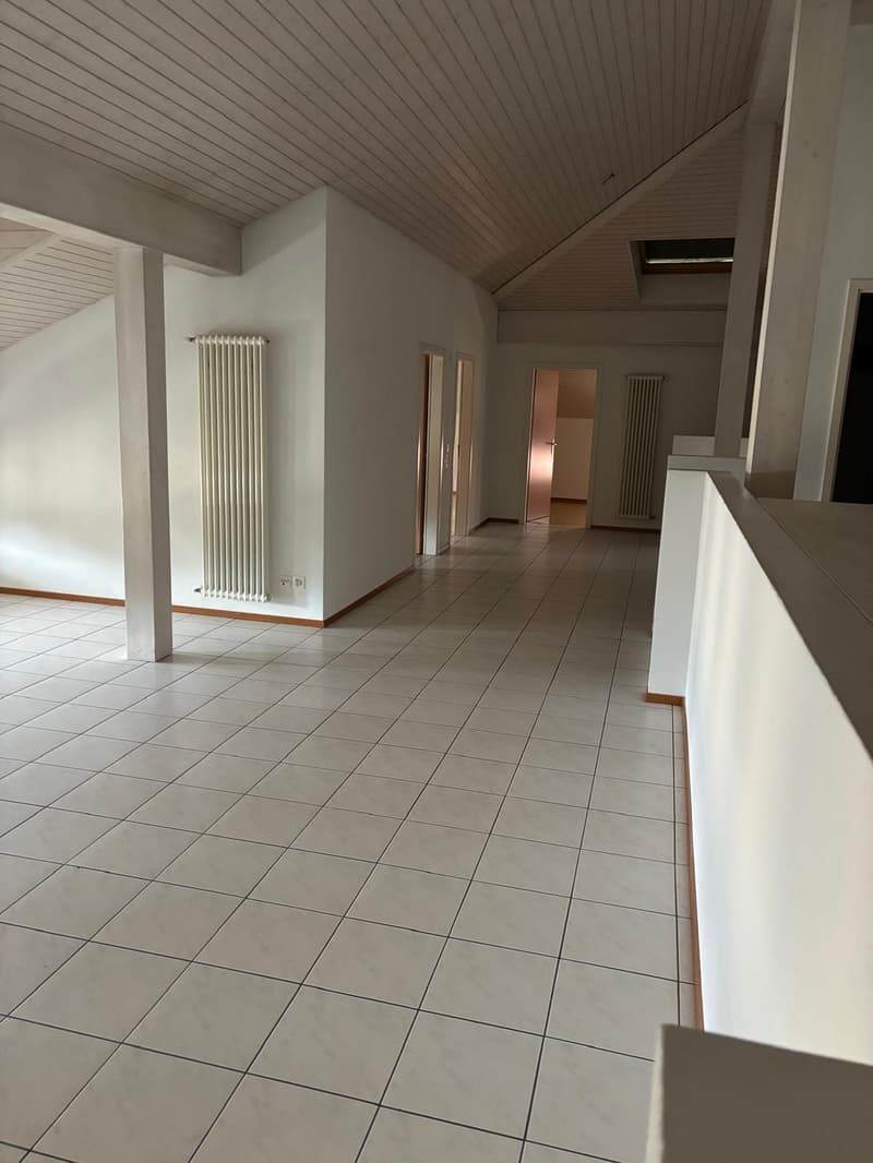The image shows a spacious interior with a tiled floor and a vaulted ceiling with wooden paneling. The room has white walls and radiators along the walls. The space appears to be a hallway or entryway leading to other rooms.