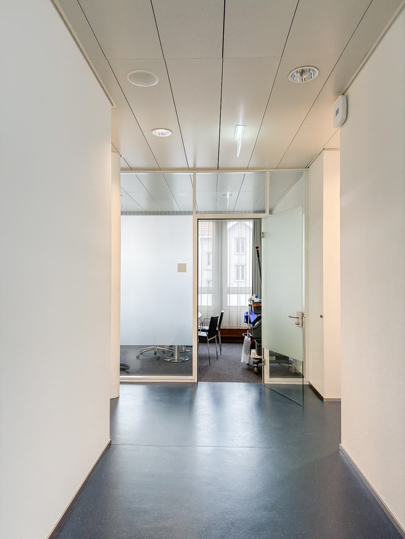 Office corridor, gray and white walls, glass partitions, ceiling lights, office furniture and chairs.